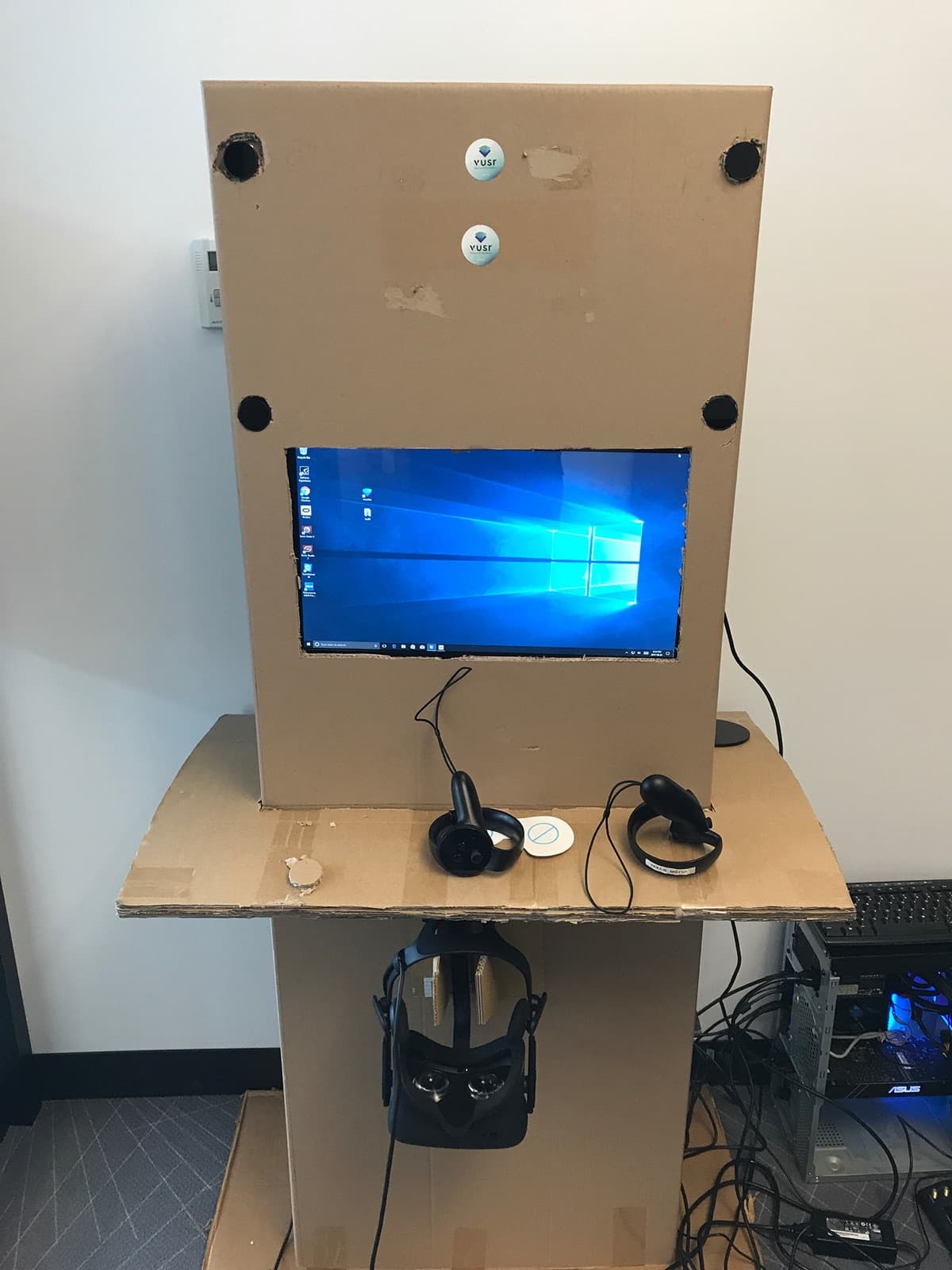 The first Vusr kiosk prototype: a cardboard tower with a monitor showing Windows cut into it, Touch controllers on a cardboard shelf, an Oculus Rift hanging beneath, and a gaming PC on the floor beside it