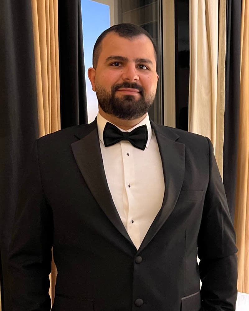 Khaled Shoukry standing by a window in a black tuxedo and bow tie, smiling at the camera